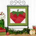  The completed June project depicting a strawberry in red, white, and green fabrics, hung on a craft display stand in front of a white paneled wall, and staged with coordinating flowers and décor.