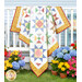 The completed vibrant patchwork Sunshine Day Quilt, colored in prints from the 30s Playtime collection by Moda Fabrics, draped on a white porch railing, surrounded by colorful flowers and a blue house.