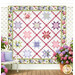 The completed Starry Lattice Quilt, colored in pink, purple, green, periwinkle, and white fabrics from the Hydrangeas in the Garden collection by Jennifer Bosworth for Riley Blake Designs, hung on a white paneled wall, paired with white lattices and full bloom hydrangea flowers.