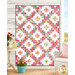 The completed Layer Cake Ladder Quilt, colored in the vibrant pink and turquoise fabrics of the Zinnias collection contrasted with white background fabric. The quilt is hanging on a white shiplap wall, with coordinating farmhouse décor and floral bouquets. There is a gold Shabby Fabrics logo in the bottom right corner.
