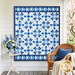 The completed Fly Over Quilt colored in blue and white fabrics from the Amalfi coast collection by White Owl Textiles. The quilt is hanging outdoors, on a white wall with coordinating blue and white décor surrounding it.