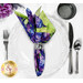 A top down shot of the napkin with the purple side out, showcasing the floral print; the napkin is staged with a white plate, silverware, and matching cup.