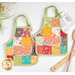 The completed Apron Hot Pads colored in red, teal, orange, and green fabrics from the In The Garden collection, staged on a white countertop with coordinating flowers and kitchen utensils; a recipe pokes out from inside one hot pad.