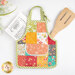 A single apron hot pad staged on a. white table while a recipe and a wooden spoon poke out of the pocket. 