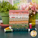 A stack of the fabrics included in the Garden Glam collection, with flowers and a jar and a flower pot and a plant and thread and a window.