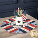 The completed Easy Patriotic Table Topper Kits finished in American themed fabrics from the Founding Glory collection by Wilmington Prints, set on a wooden table with a white jar filled with red, white, and blue florals. 