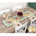 The completed table runner made with log cabin blocks of plum purple, ocher, and teal floral fabrics from the Renew collection, laying in the center of a wooden dining table with white chairs and four place settings at each seat with matching cloth napkins.