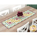 The completed table runner made with log cabin blocks of plum purple, ocher, and teal floral fabrics from the Renew collection, laying in the center of a wooden dining table with white chairs.