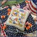 A decorative stitched pillow featuring houses, trees, and flags, staged with sewing supplies and fabric.