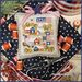 A decorative stitched pillow featuring houses, trees, and flags, staged with sewing supplies and fabric.