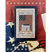 finished cross stitch project framed featuring a flag, white colonial house, and the words "broad stripes, bright stars" staged with a flag and decor