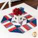 The completed Easy Patriotic Table Topper colored in patriotic themed fabrics from the Celebrate The USA collection by Benartex, staged on a white table with a coordinating white vase with red and blue flowers.