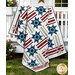 The completed Hometown Honor quilt, a geometric stripe and star design colored in gray, white, red, and blue batik fabrics from the Missing You collection from Island Batik, artfully draped over a white porch railing onto a green lawn with flowers.