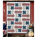 The completed Hometown Honor quilt, a geometric stripe and star design colored in gray, white, red, and blue batik fabrics from the Missing You collection from Island Batik, staged on a red barn wall with coordinating décor.