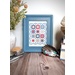 A blue-framed cross-stitch artwork sits on a wooden table, surrounded by a vase and buttons.
