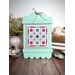 A mint-framed cross-stitch artwork sits on a wooden table, surrounded by a vase and buttons.