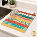 The completed Dry Ideas drying mat, colored in vibrant prints from the Desert Dreams collection by Maywood Studio, staged on a marble kitchen counter and holding two glasses.
