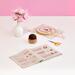 Open booklet of various pink stitches on a pink background with scissors, pin cushion, flowers, and a hoop.
