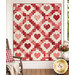 The completed Heartsy Quilt, colored in pink, red, and cream fabrics from the Petals and Polka Dots collection by Moda Fabrics, staged on a white porch wall with coordinating furniture and flowers.