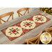 The completed Any 12 Will Do Table Runner, colored in cream, green, and red metallic fabrics from the Merry Manor Metallic 2025 collection by Moda Fabrics, staged on a light wooden table with four chairs and a decorated Christmas tree peeking into frame from the upper right corner.