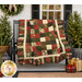 The completed Holiday Charm Quilt, colored in cream, green, and red fabrics from the Merry Manor Metallic 2025 collection, artfully draped over a black bench on a white porch, staged with coordinating plants and décor.