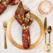 A top down shot of the napkin with the red fabric facing out, staged on a coordinating gold table setting on a wood table.