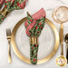 A top down shot of the napkin with the green holly fabric facing out, staged on a coordinating gold table setting on a white marbled table.