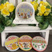 The fully assembled and painted Fleur Mini Round Cross Stitch Frame, staged on a white pedestal framed by yellow flowers, displaying four festive cross-stitch projects.