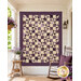 The completed Playing Favorites Quilt colored in rich cream and purple fabrics from the Plumberry Farm collection by Marcus Fabrics, hung on a cream porch wall and staged with coordinating décor. 