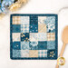 The completed Fusible Grid Hot Pad colored in cream and blue fabrics from the Special Delivery collection by Andover Fabrics, staged on a white marbled table where a wooden spoon and coordinating flowers peek into frame.