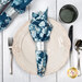 A top down shot of the napkin with the dark blue floral fabric facing out, staged on a coordinating cream and silver table setting on a wood table.