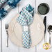 A top down shot of the napkin with the gingham fabric facing out, staged on a coordinating cream and silver table setting on a wood table.