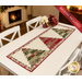 The completed Tree Farm Table Runner colored in cream, red, green, and black fabrics from the Candlelight Christmas collection by Moda Fabrics, staged on a white table with coordinating décor.