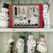 Finished Peaceful Snow cross stitch surrounded by vintage winter dolls on a white painted shelf.