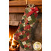 The completed Fast Fusible Patchwork Large Stocking, colored in cream, green, and red metallic fabrics from the Merry Manor Metallic 2025 collection by Moda Fabrics, hung on a warm wooden mantle of a lit fireplace.