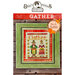 Front cover of the finished Gather cross stitch pattern with two pilgrims holding Thanksgiving food in a green frame laid on wooden planks.