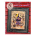 Front cover of the finished Haunted Forrest cross stitch pattern of a haunted house in a forest with Halloween motifs inside of a black wood frame hung on a red wooden backdrop.