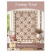 The front cover of the Morning Blush pattern showing the completed quilt colored in pastel pink, brown, cream, and red fabrics, staged on an off white porch wall with coordinating furniture and décor.