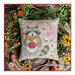Finished The Little Raccoon cross stitch pattern on a linen decorative pillow on a wooden stump surrounded by flowers, thread, and sewing scissors.