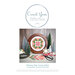 Front cover of the Whimsy Star cross stitch pattern showing the finished product in a wooden circle hoop next to frosty prop trees, a snowflake, the Moda Poinsettia and Pear line, and a tub of buttons.
