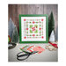 Finished Wreath and Pine cross stitch pattern in a green square frame surrounded by frosted pine trees, a snowflake, Moda's Poinsetta and Pear fabrics, and a tub of buttons.
