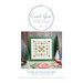 Front cover of the finished Wreath and Pine cross stitch pattern in a green square frame surrounded by frosted pine trees, a snowflake, Moda's Poinsetta and Pear fabrics, and a tub of buttons.