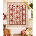The completed Sunday Picnic Quilt, colored in warm fabrics from the Hearth & Home collection by Henry Glass Fabrics, staged on an off-white porch wall and surrounded by coordinating furniture and décor.