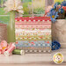 A stack of the fabrics included in the Meadowbrook collection with flowers and vases and thread and a window.