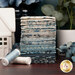 A stack of the fabrics included in the La Maison Bleu collection, with plants and flowers and a ceramic house and thread.