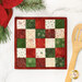 The completed Fusible Grid Hot Pad, colored in red, green, and cream fabrics from the Christmastime collection by Maywood Studio, staged on a white marble kitchen counter with coordinating decor.