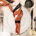 A top down shot of the napkin with the orange and white cat ghost print side out, showcasing the geometric print; the napkin is staged with a cream plate and matching table setting.