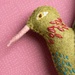 Close-up of the green hummingbird with stitched details on a pink background.