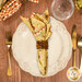 A top down shot of the napkin with the yellow floral fabric facing out, staged on a coordinating cream table setting on a wood table.
