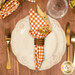 A top down shot of the napkin with the orange and white gingham fabric facing out, staged on a coordinating cream table setting on a wood table.